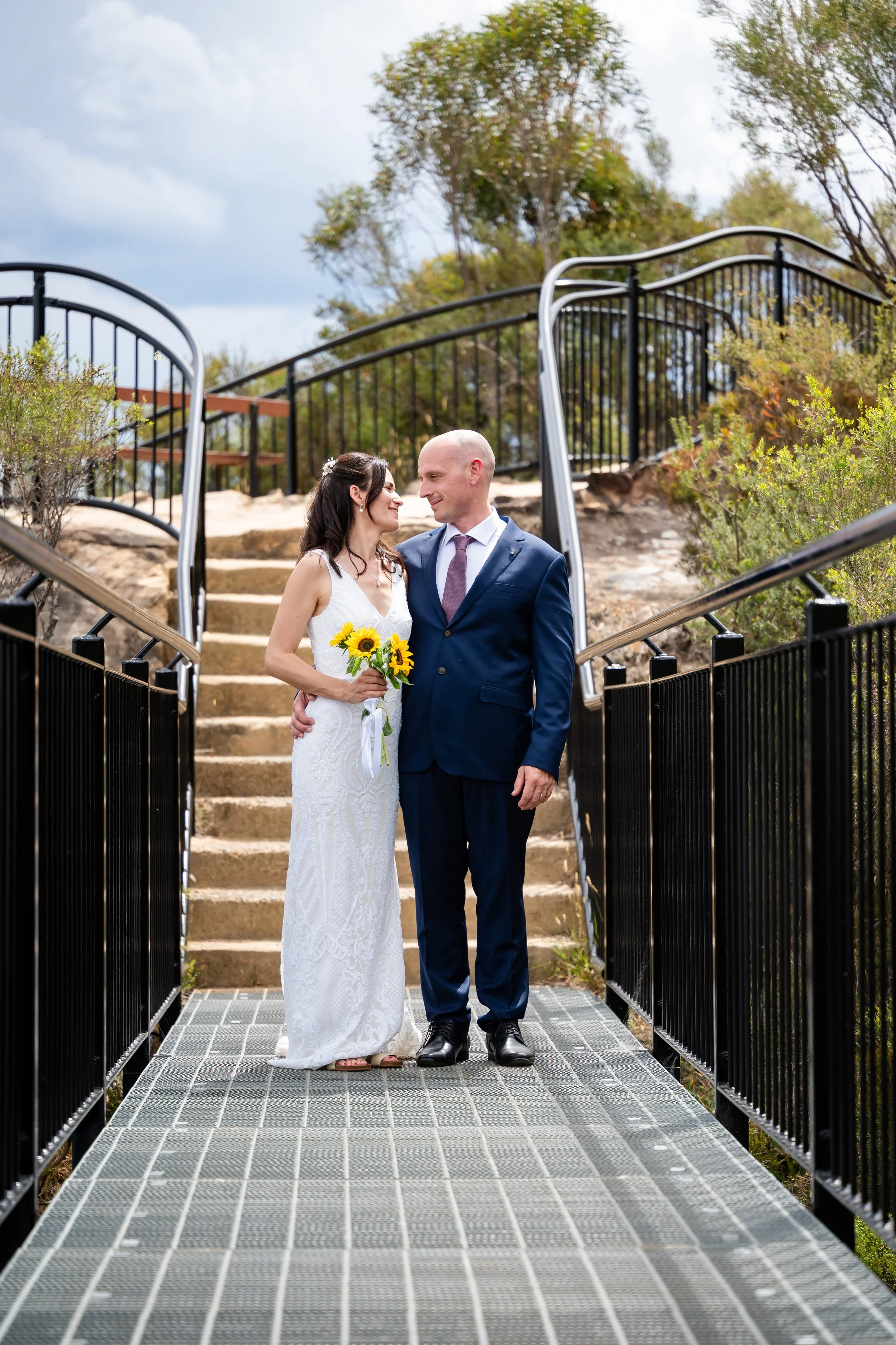 A bride and groom standing on a metal bridge, gazing lovingly at each other, with the bride holding a bouquet of sunflowers. The bride is wearing a white dress and the groom is dressed in a navy suit. There are stairs and greenery in the background.