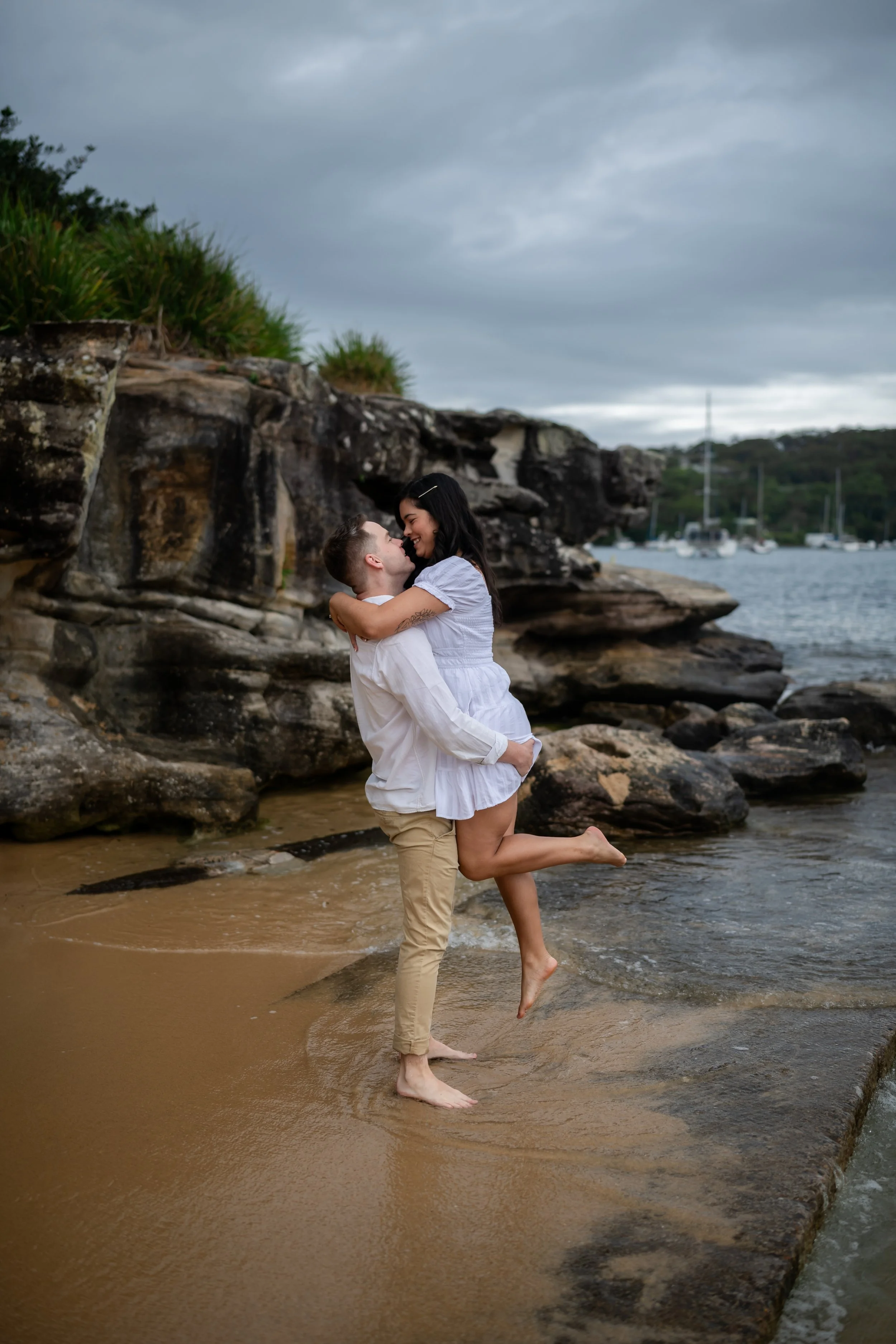 A couple hugging and smiling at each other on a beach with rocks and boats in the background.