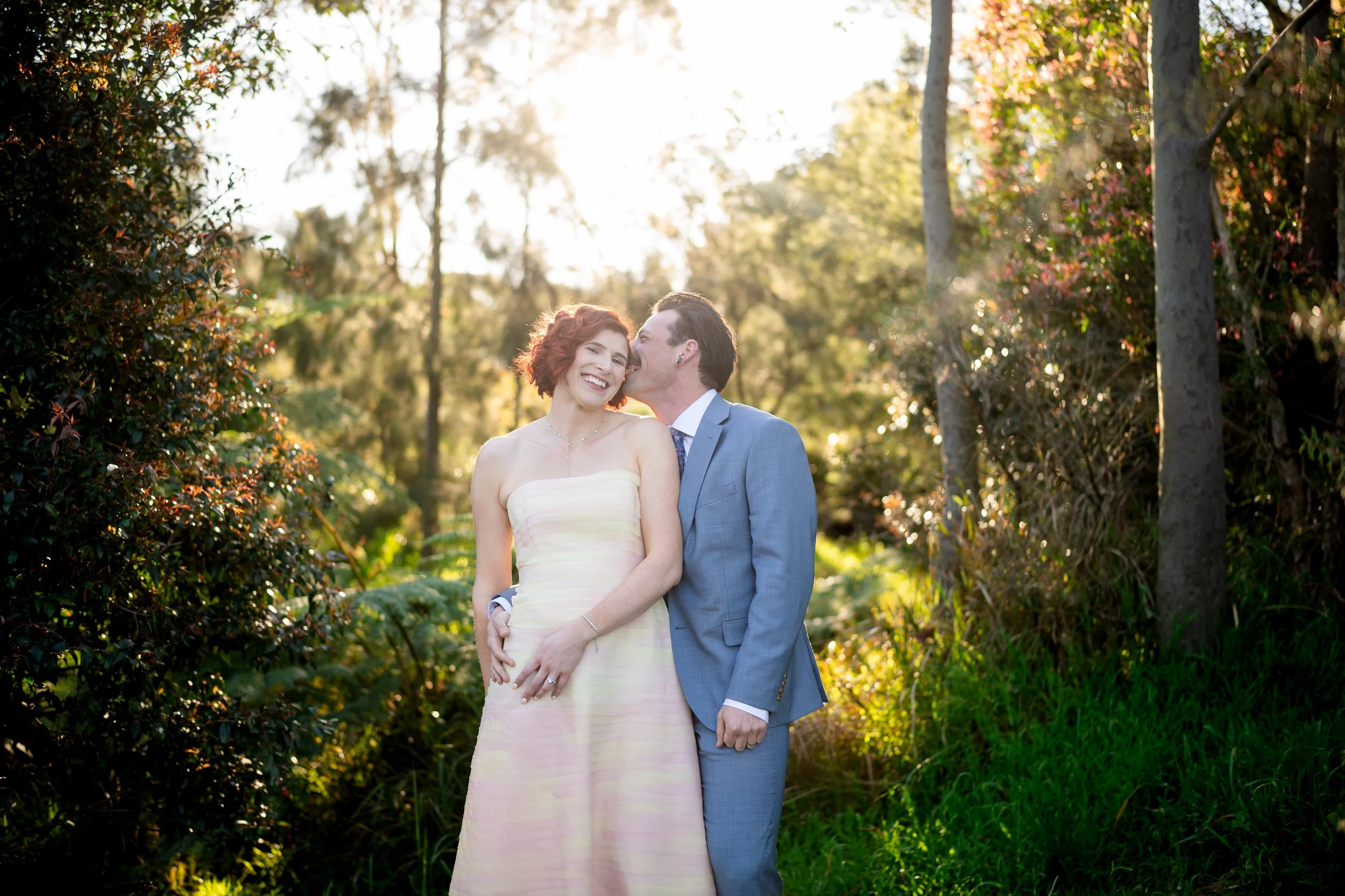 A couple dressed in wedding attire standing in a wooded outdoor setting, with the man kissing the woman on the cheek, both smiling, during sunset.