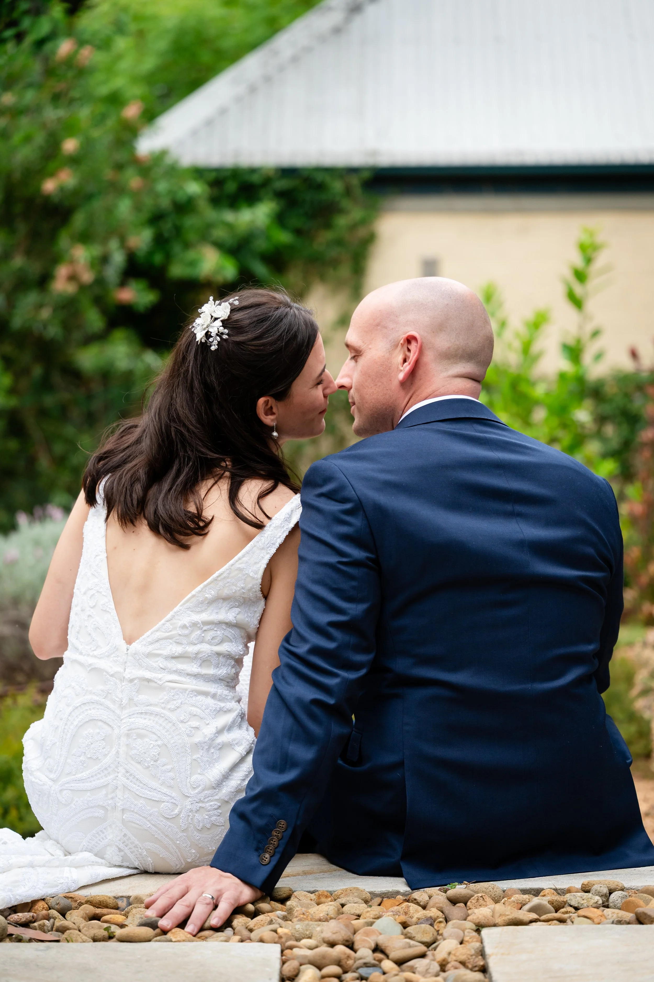 A bride and groom sitting outdoors on a stone and wood surface, leaning close with their noses touching, in a garden setting with greenery and a building in the background.