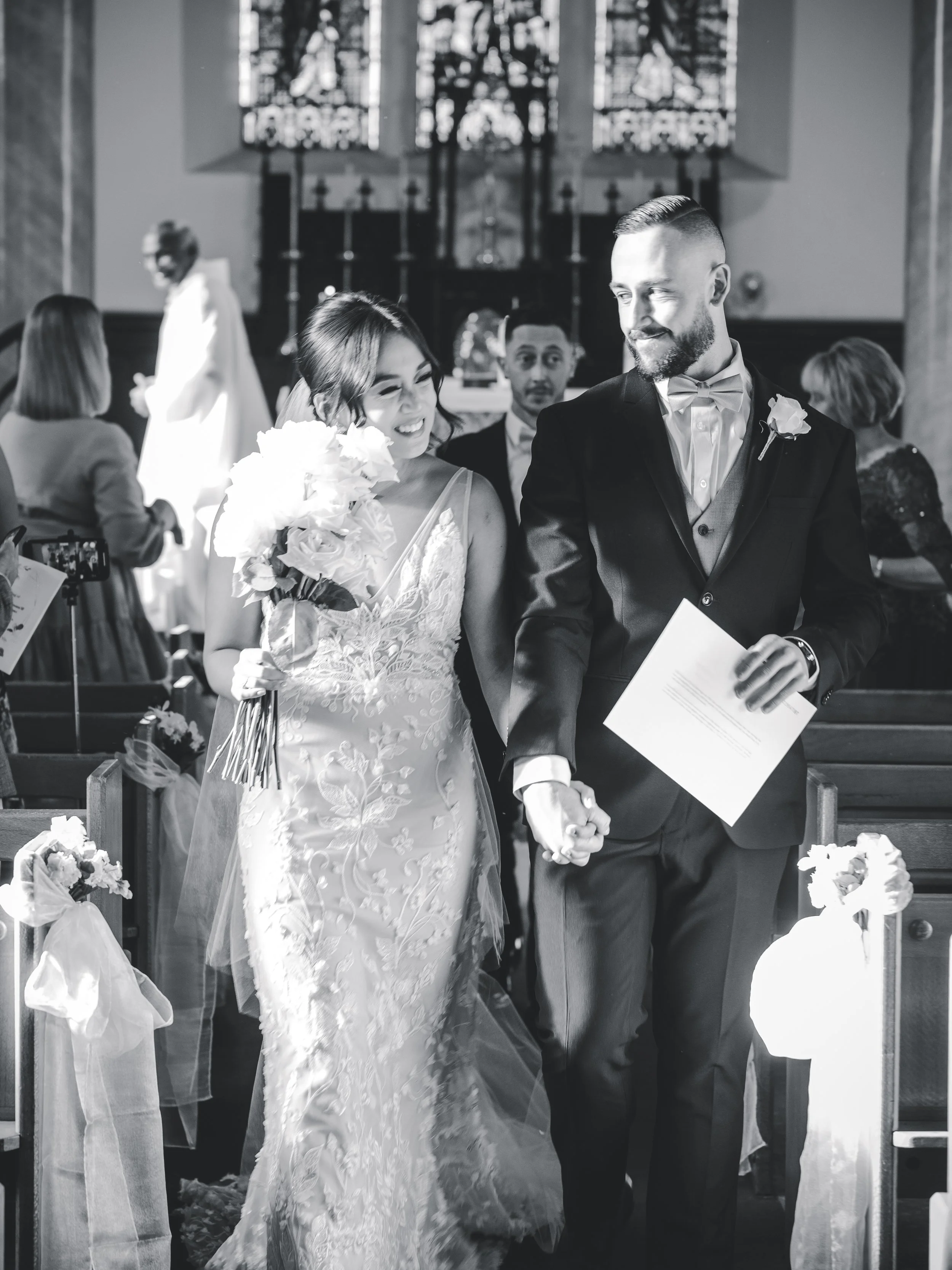 Black and white photo of a bride and groom holding hands during their wedding ceremony inside a church, with guests and a priest in the background.