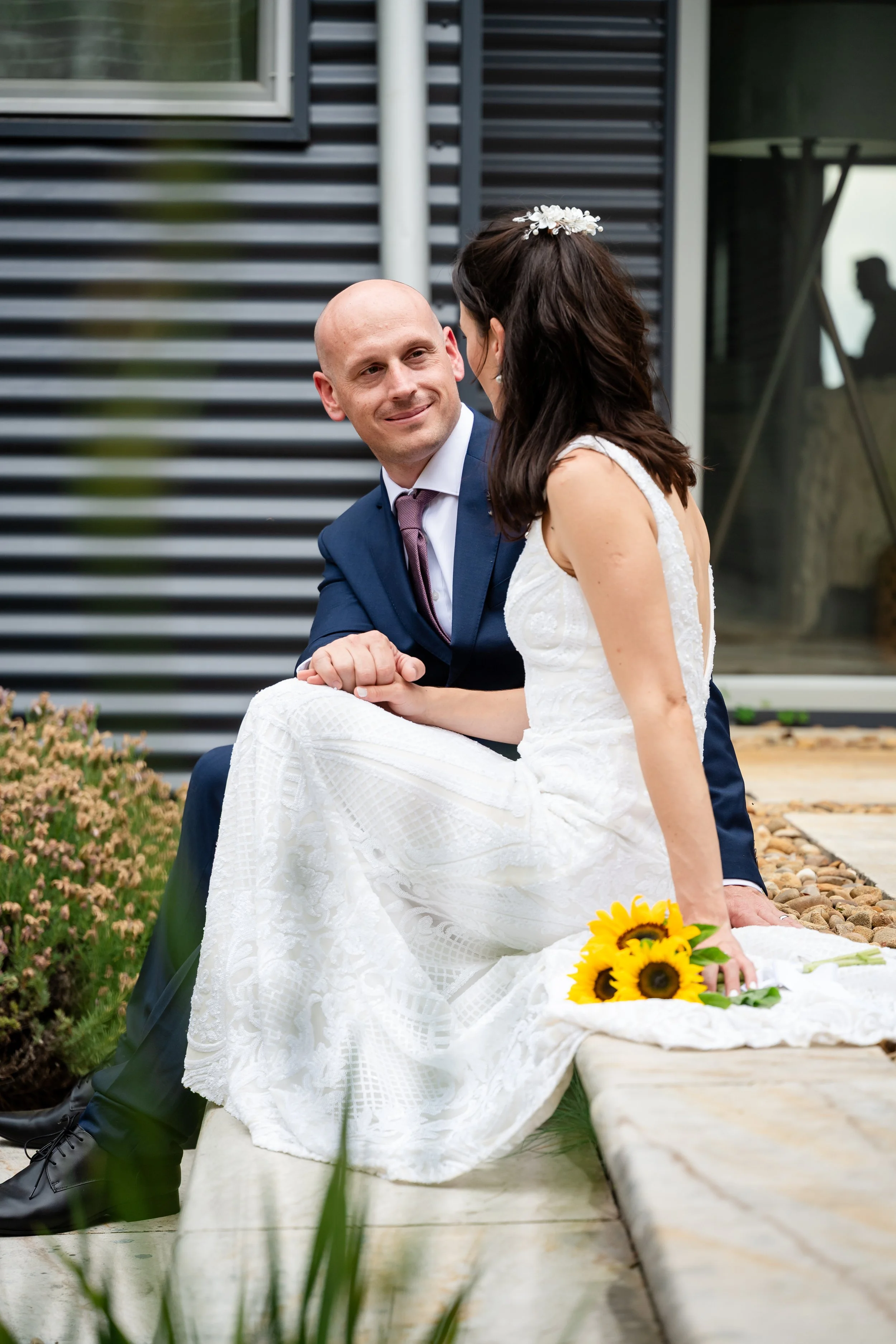 A newlywed couple sitting outside, the man in a navy suit and the woman in a white wedding dress, holding hands and looking at each other, with a bouquet of sunflowers on the ground beside her.