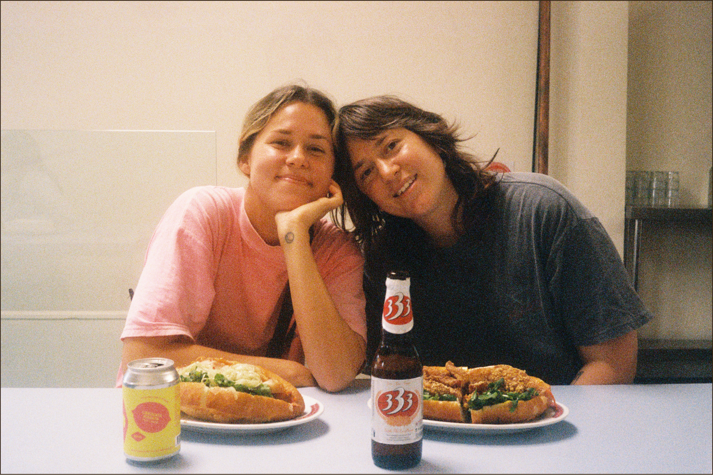 Emma and Lisa of Soft Power sitting at a table with pizza, a can of soda, and a beer bottle, smiling and leaning on each other in an indoor setting.