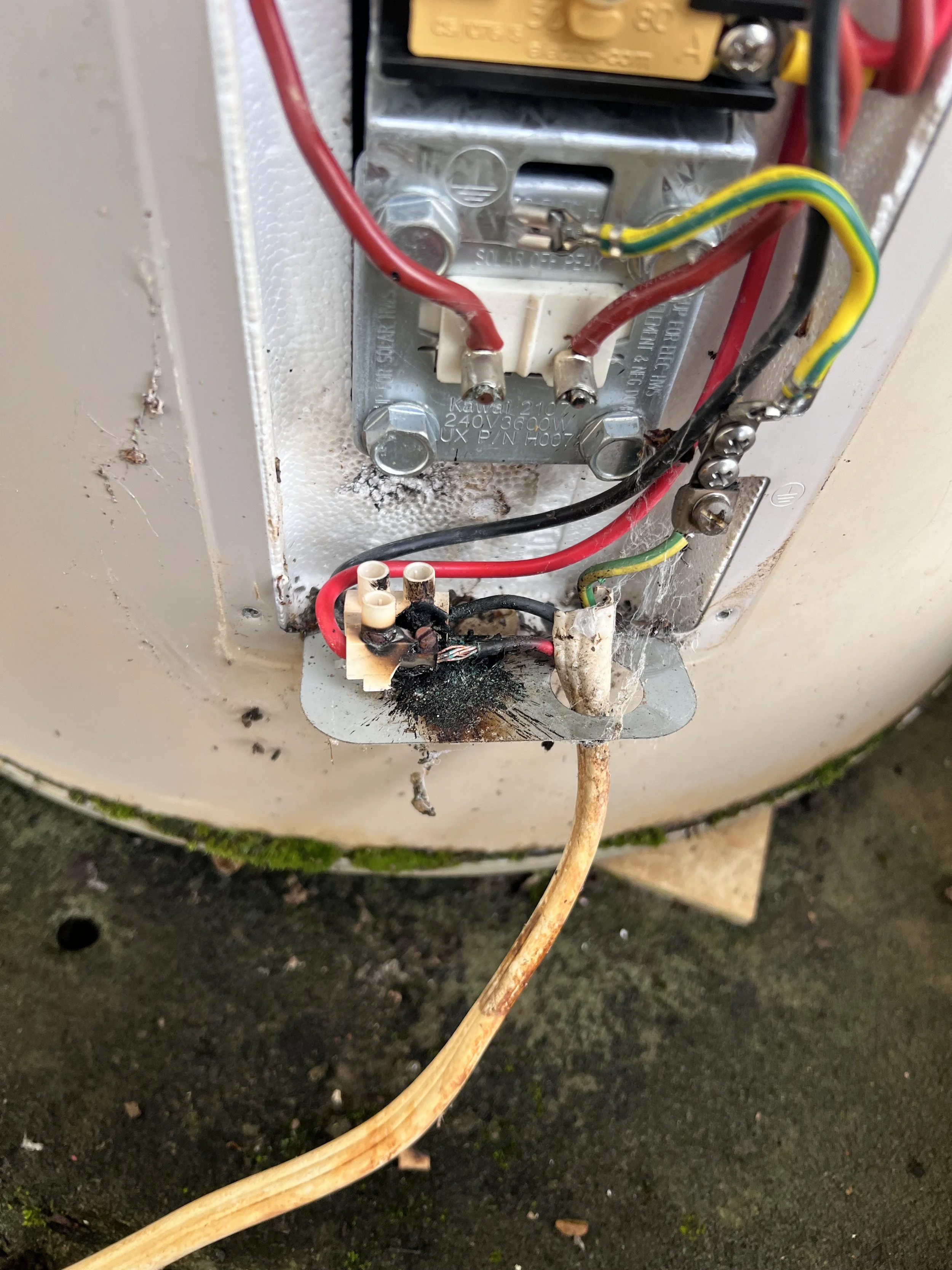 Close-up of an electrical hot water heater and faulty wiring, showing red, black, and green wires connected to components inside the box.