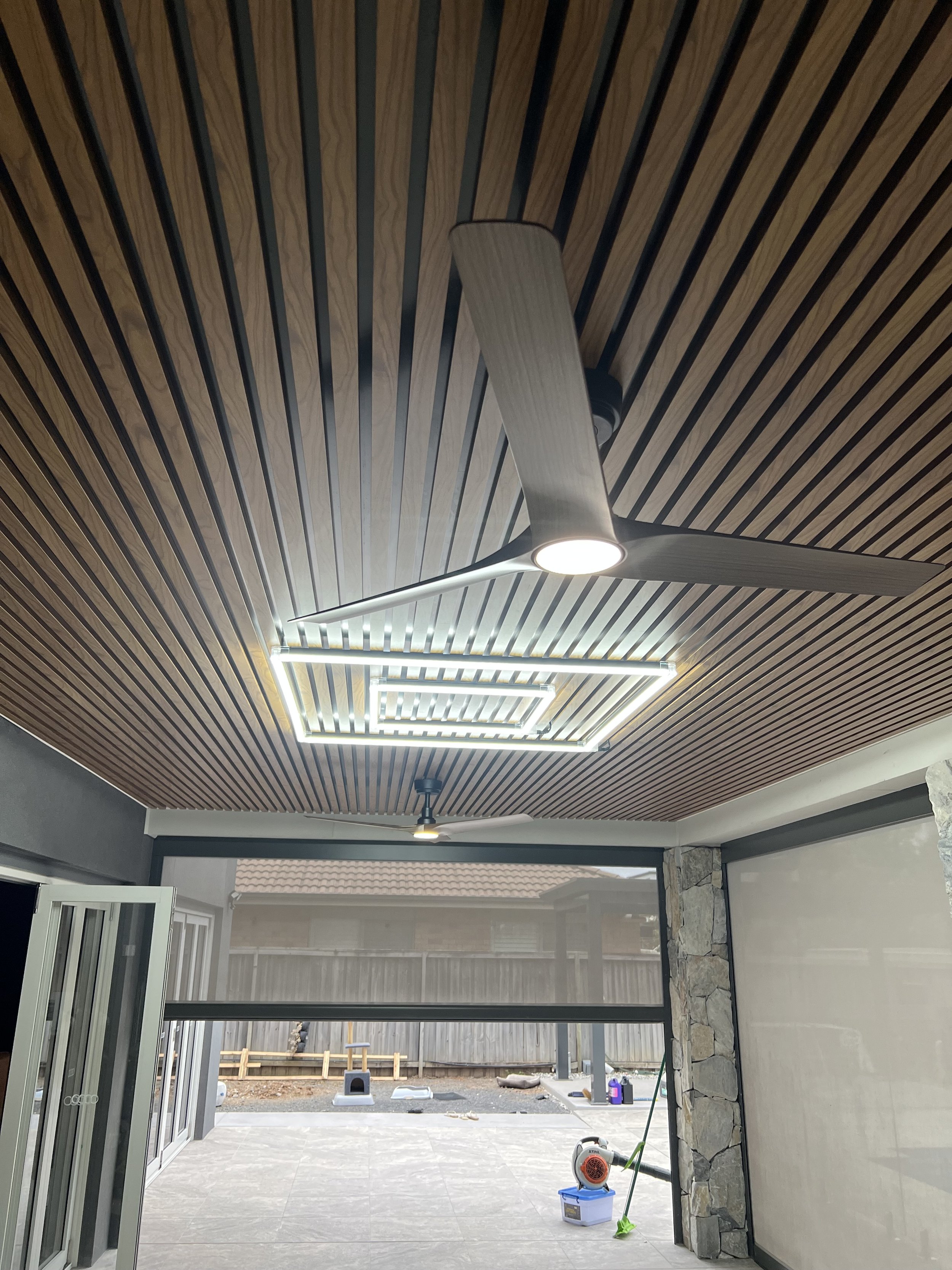 A covered patio or porch area with a wooden slat ceiling and a ceiling fan with a light. And accent lighting on the roof.