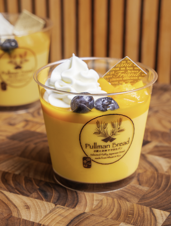 A orange mango pudding dessert in a clear cup topped with whipped cream and grapes, with a Pullman Bread logo on it, placed on a wooden surface with a blurred background.