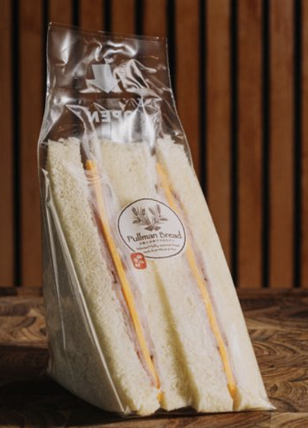 Sandwiches in a clear plastic bag with pulled bread branding, placed on a wooden surface with a wooden panel background.