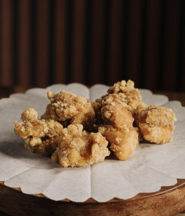 Six pieces of fried chicken on a piece of parchment paper, set on a wooden plate against a dark background.