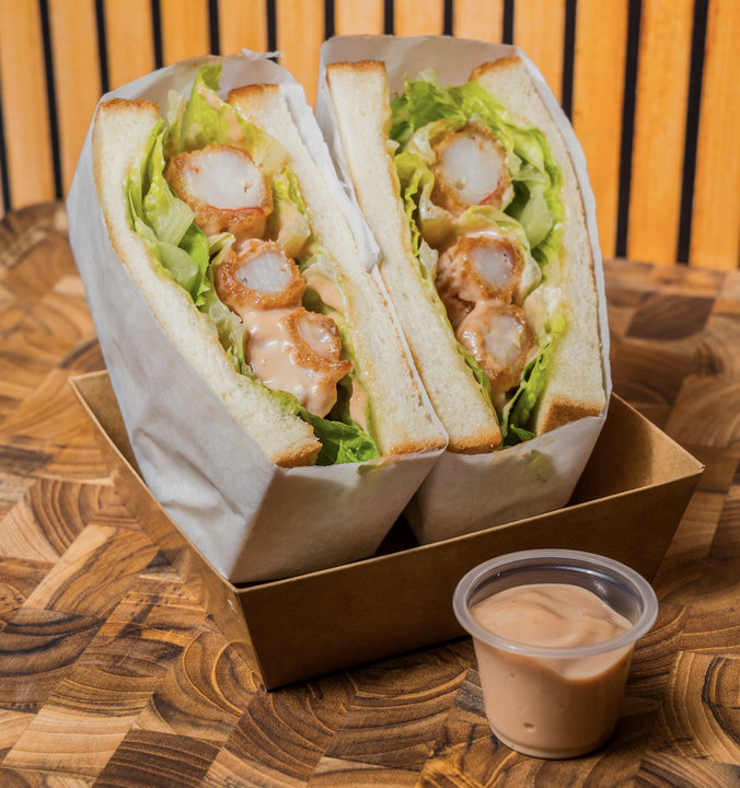 Two shrimp sandwich halves with lettuce and sauce, served with a side of dipping sauce in a small plastic cup, placed in a brown paper tray on a wooden table.