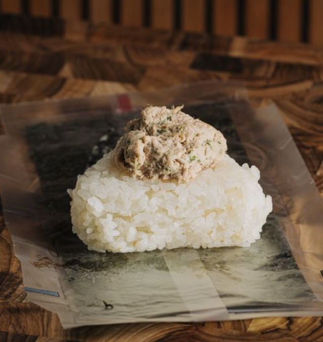 A rice ball topped with a dollop of creamy, chunky fish or seafood mixture, placed on a piece of printed paper on a wooden surface.