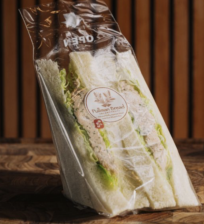 Pre-packaged sandwich in clear plastic bag with Pullman Bread label and lettuce, on a wooden surface