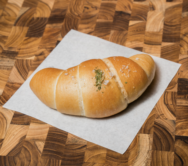 A butter roll, topped with herbs and salt, resting on a piece of white paper on a wooden table.