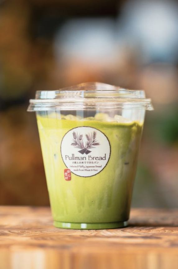 A plastic cup filled with green matcha latte iced beverage, featuring a Pullman Bread logo with ingredients listed, placed on a wooden surface.