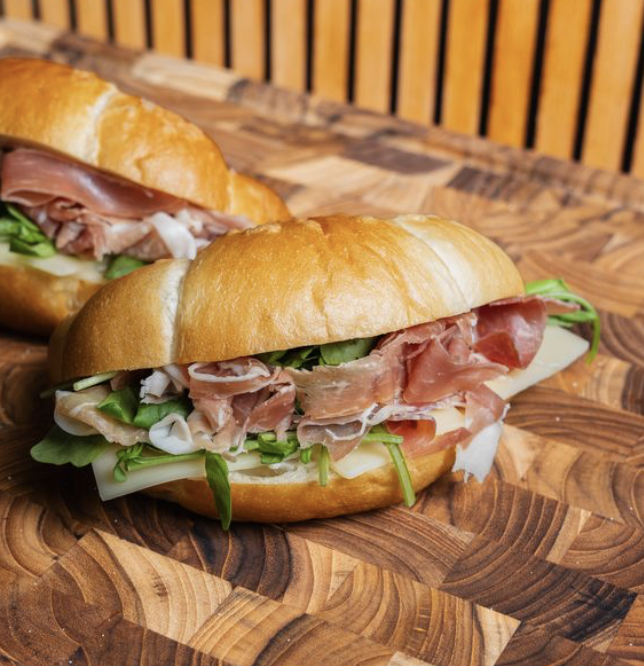 Two butter roll sandwiches with slices of ham, arugla, and cheese on a wooden surface.