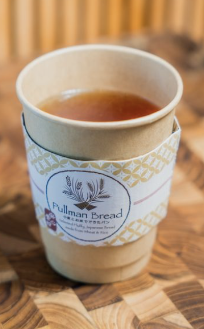 A paper cup filled with tea, wrapped with a sleeve from Pullman Bread, placed on a wooden table.