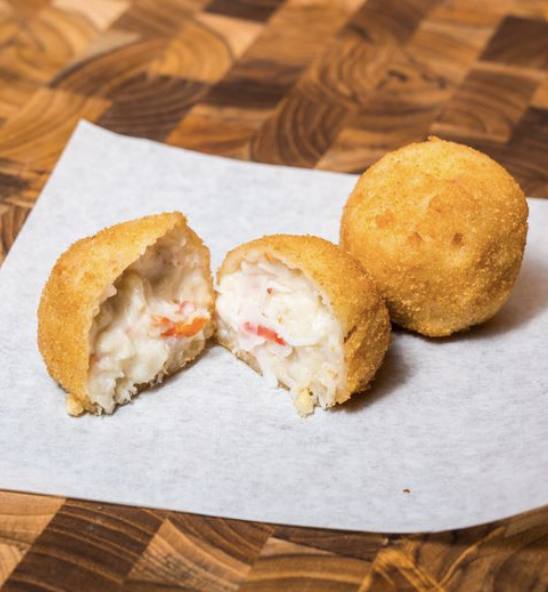 Two fried crab croquette balls, one halved to show creamy filling with small bits of red and orange, on white paper on a wooden surface.