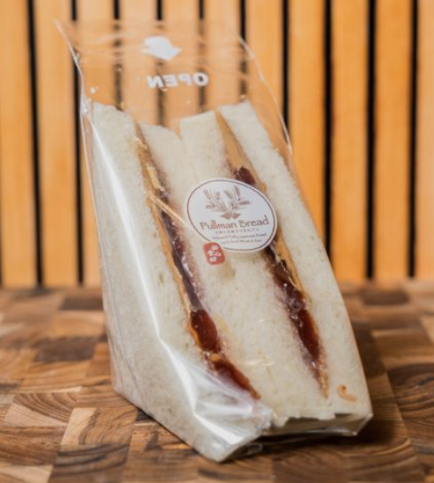 A pre-packaged Pullman bread sandwich with visible jam filling inside a clear plastic bag, placed on a wooden surface with a wooden slat background.
