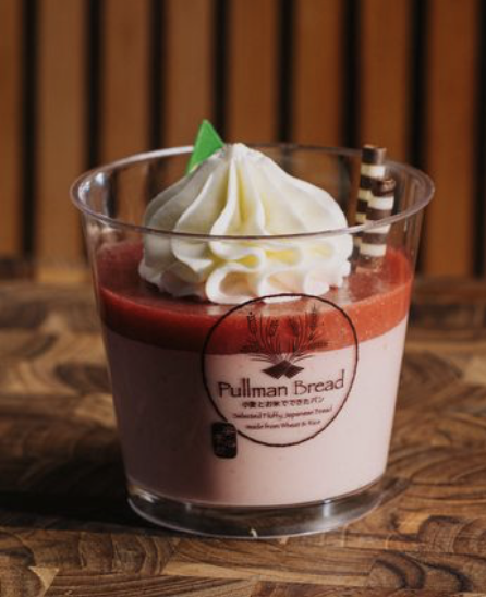 A dessert in a clear glass cup with layers of red and pink strawberry flavor, topped with whipped cream and a small green garnish, on a wooden surface.
