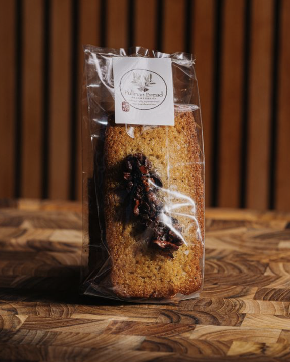 A packaged financier with a label that reads 'Pullman Bread' and a cluster of dark cacao nib on the top, placed on a wooden surface with a background of vertical wooden panels.