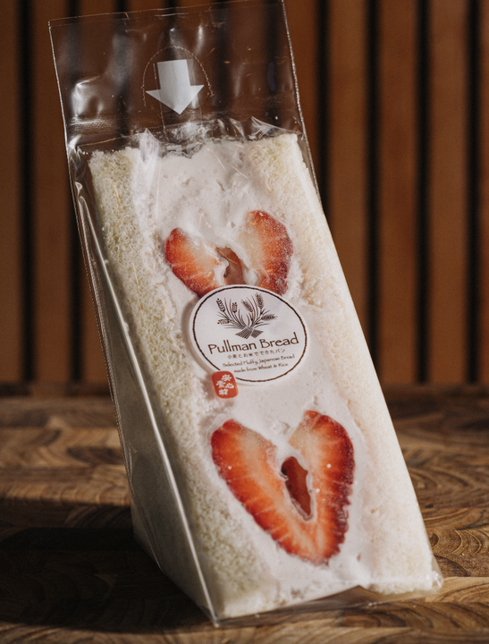 Prepackaged strawberry and cream sandwich in clear plastic wrapping with Pullman Bread label, on wooden surface with vertical wooden slats in background.