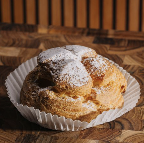 A cream puff pastry dusted with powdered sugar placed in a white paper liner on a wooden surface.