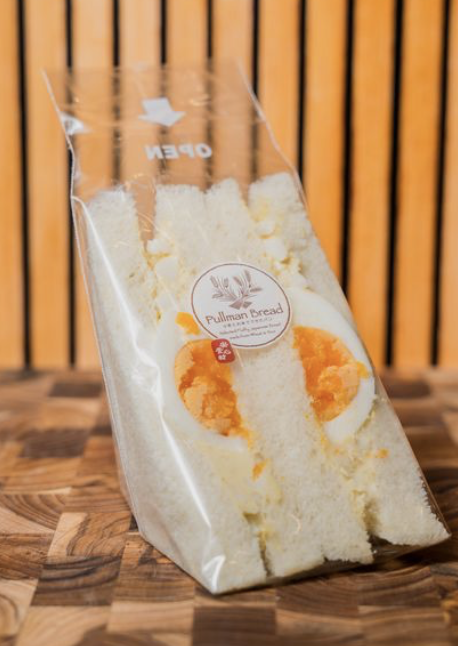 A clear plastic bag containing a bread sandwich with egg filling, labeled Pullman Bread, placed on a wooden surface with a wooden slat background.