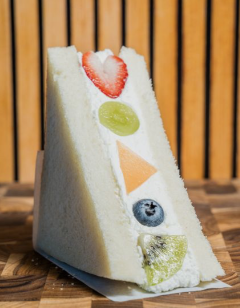 A slice of white bread sandwich with fruit decorations including a strawberry heart, green grape, orange triangle, blueberry, and kiwi, arranged vertically on the side.