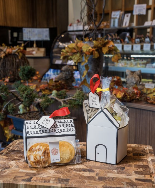 Handmade gift boxes with cookies and baked goods on a wooden table in a store decorated with autumn leaves and plants.