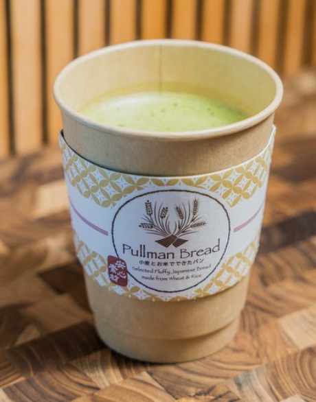 A disposable coffee cup filled with hot matcha latte with a label from Pullman Bread, placed on a wooden surface with a blurred wooden background.