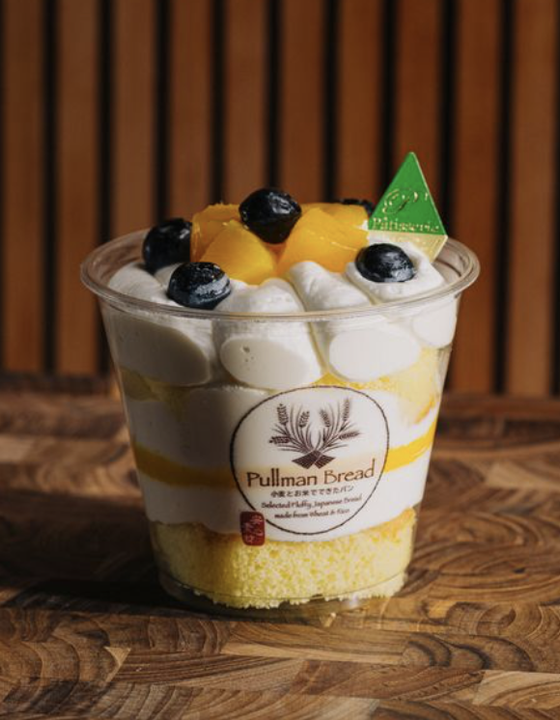 Dessert parfait with layers of cream, yellow cake, blueberries, mango topping, labeled 'Pullman Bread' in a clear cup on a wooden surface.
