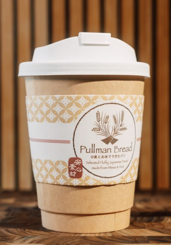 Disposable coffee cup with a white lid and a paper sleeve filled with hot coffee, labeled Pullman Bread, featuring Japanese text and an illustration of wheat on a wooden surface with a wooden slat background.