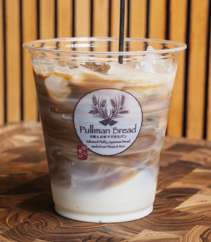 A plastic cup filled with iced coffee featuring swirls of milk and coffee, and a black straw. The cup has a Pullman Bread logo sticker with Japanese characters and a wheat design. The cup is placed on a wooden table against a wooden panel background.