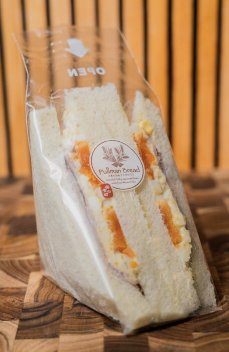 A packaged sandwich with white bread and orange filling inside, labeled 'Pullman Bread,' sitting on a wooden surface with a wooden slat background.