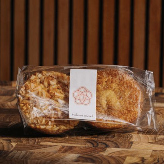 Packaged butter mochi from Pullman bread in clear plastic bag with a white label, on a wooden surface with a wooden slat background.