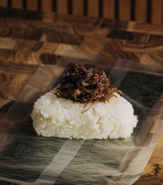An onigiri rice ball topped with cooked shredded meat, placed on seaweed paper on a wooden surface.