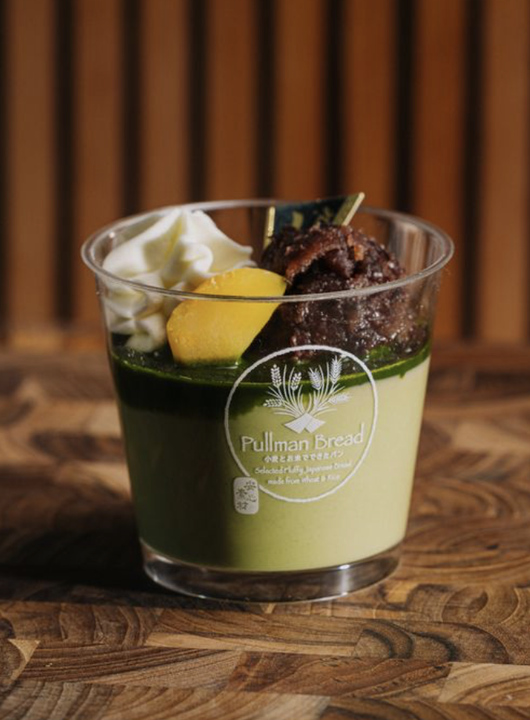 A dessert in a glass container with a green matcha layered base, topped with whipped cream, a yellow chestnut, and a brown redbean paste, set on a wooden surface with a wooden paneled background.