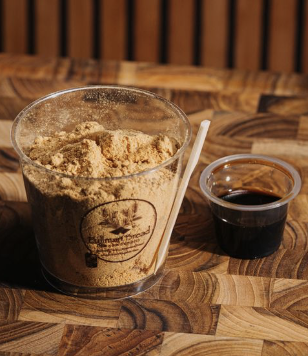 A clear plastic cup filled with powdered roasted soy and mochi, a small container of dark syrup, and a white spoon on a wooden surface.