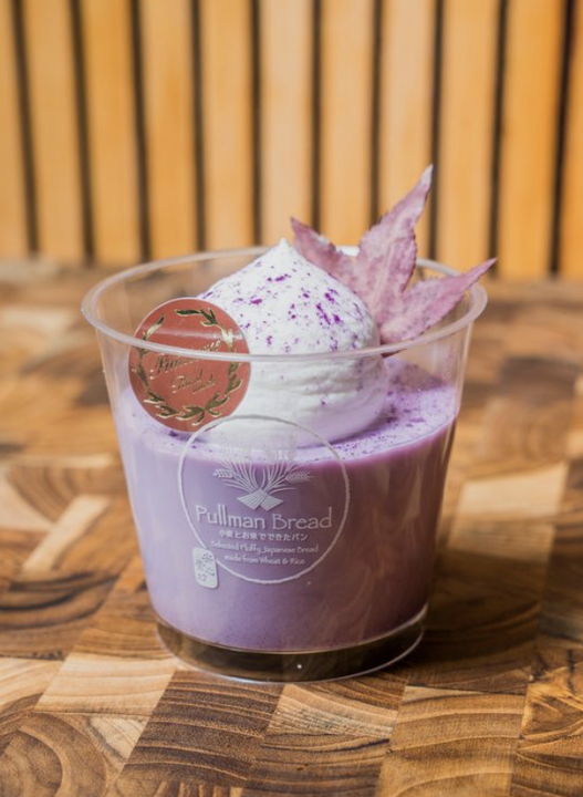 A cup of purple potato pudding topped with whipped cream, purple sprinkles, and a purple-colored decorative leaf, on a wooden surface with a wooden slatted background.