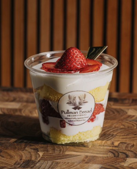 A dessert cup with layers of sponge cake, strawberries, and cream topped with a sliced strawberry, on a wooden surface with a wooden slat background.