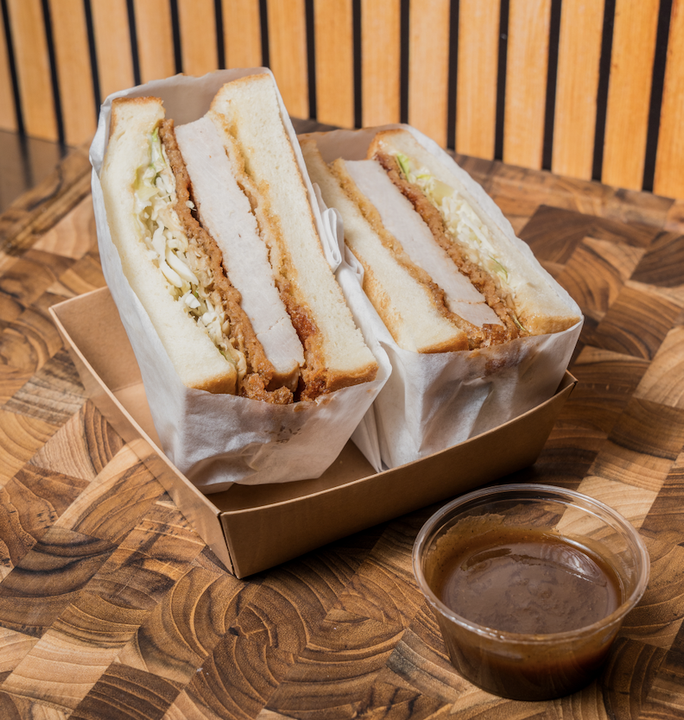 Two sandwiches with pork fillets, lettuce, and mayonnaise in white bread, served in a brown paper tray, with a small cup of dipping sauce on a wooden table.