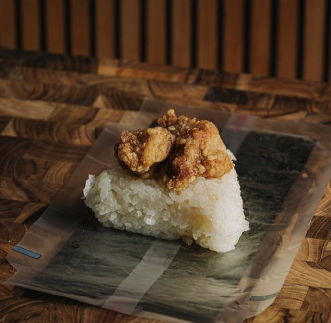 Fried chicken on top of a rice ball, placed on a sheet of seaweed, on a wooden surface.