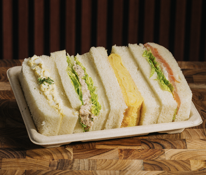 A tray of assorted sandwich slices with lettuce, egg salad, tuna salad, cheese, and smoked salmon on white bread, placed on a wooden table.