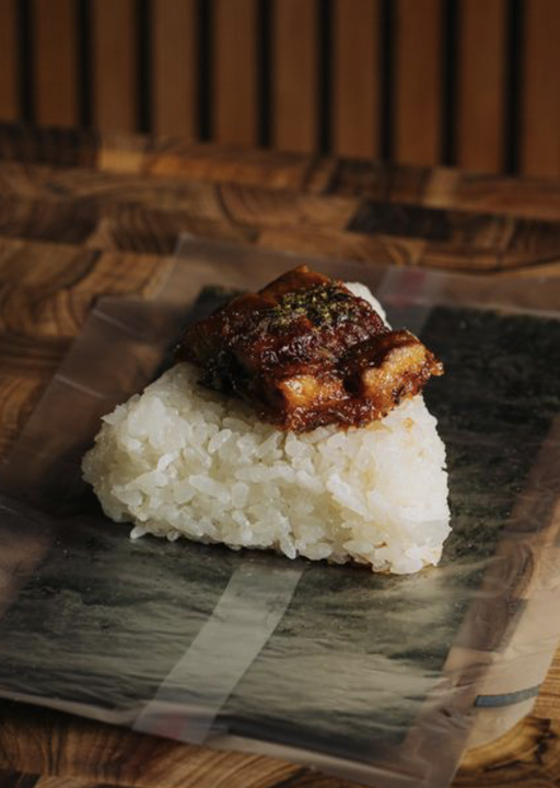 A piece of grilled eel atop a mound of white rice on a sheet of nori, served on a black plate.