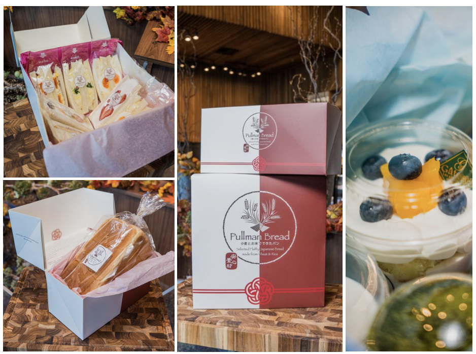 assortment of Japanese-style baked goods and desserts, including sandwiches and a fruit-topped dessert in a cup packaged in a box, displayed on a wooden surface with autumn leaves in the background.