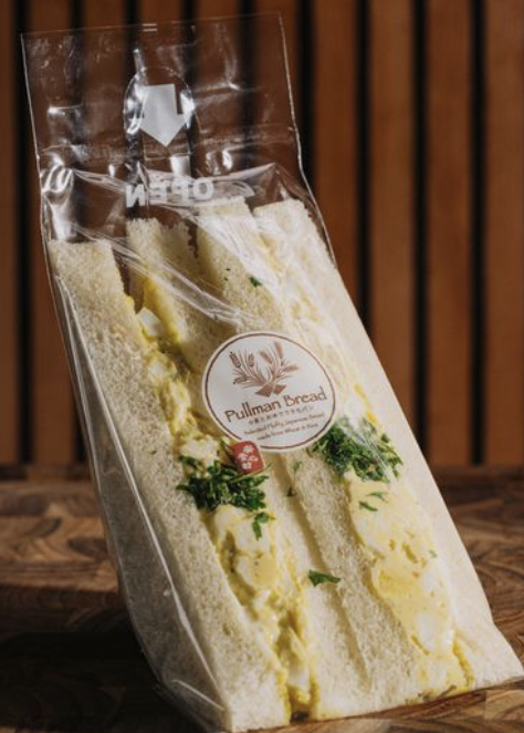 Packaged egg salad sandwich with parsley in plastic wrap on a wooden surface.