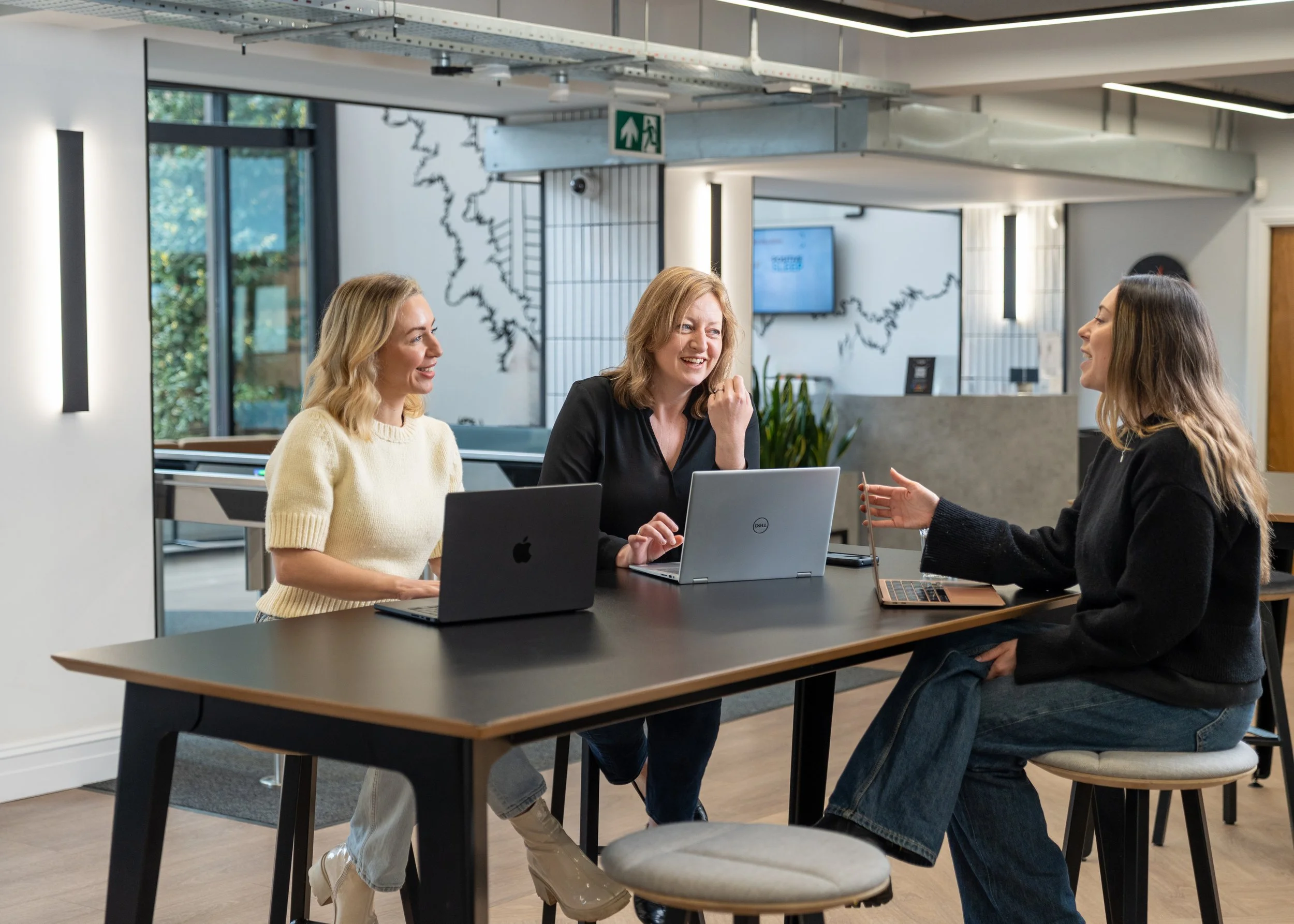 Karis Copp Media | B2B Print Industry Communications Agency in UK | Three women sitting at a conference table, engaged in conversation, with laptops in front of them in a modern office space.