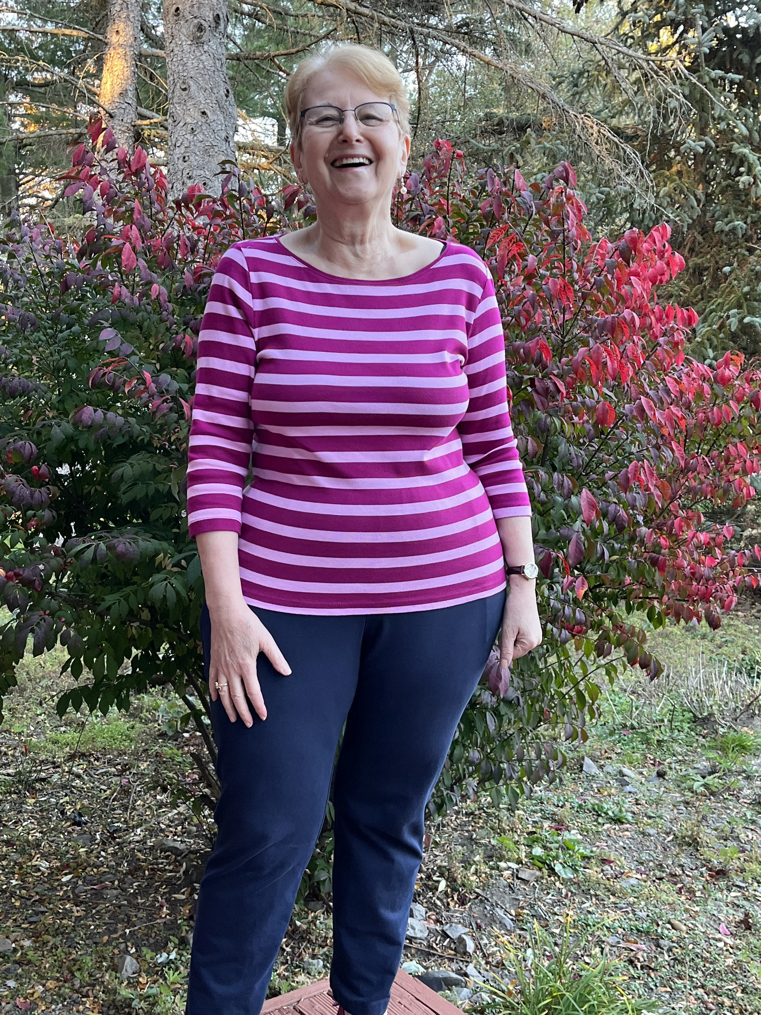 A smiling older woman with glasses and short blonde hair standing outdoors in front of a red and green shrub, wearing a pink and purple striped long-sleeve shirt and dark pants.