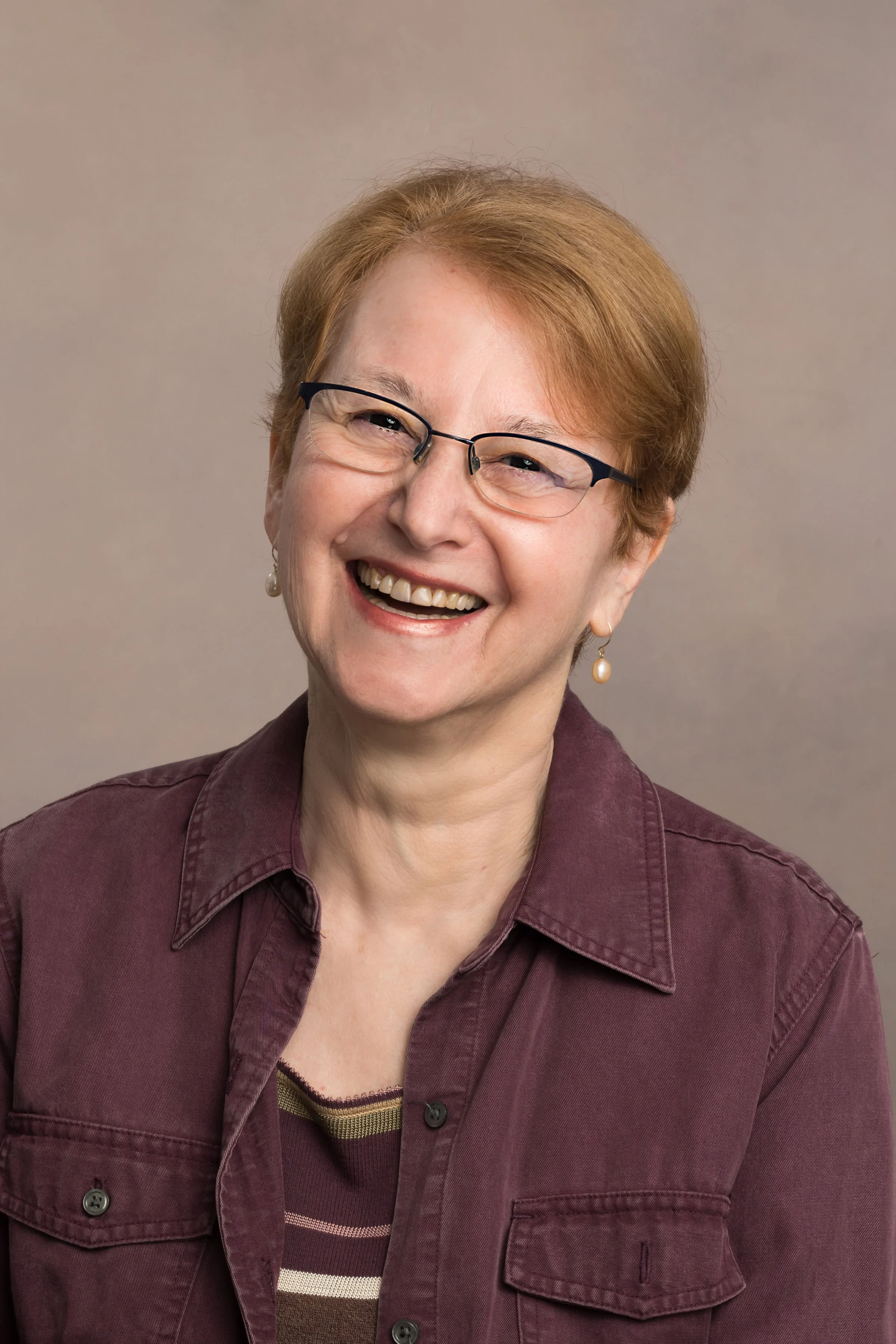 Portrait of a smiling woman with short red hair, glasses, pearl earrings, wearing a maroon shirt with a striped t-shirt underneath, against a plain background.