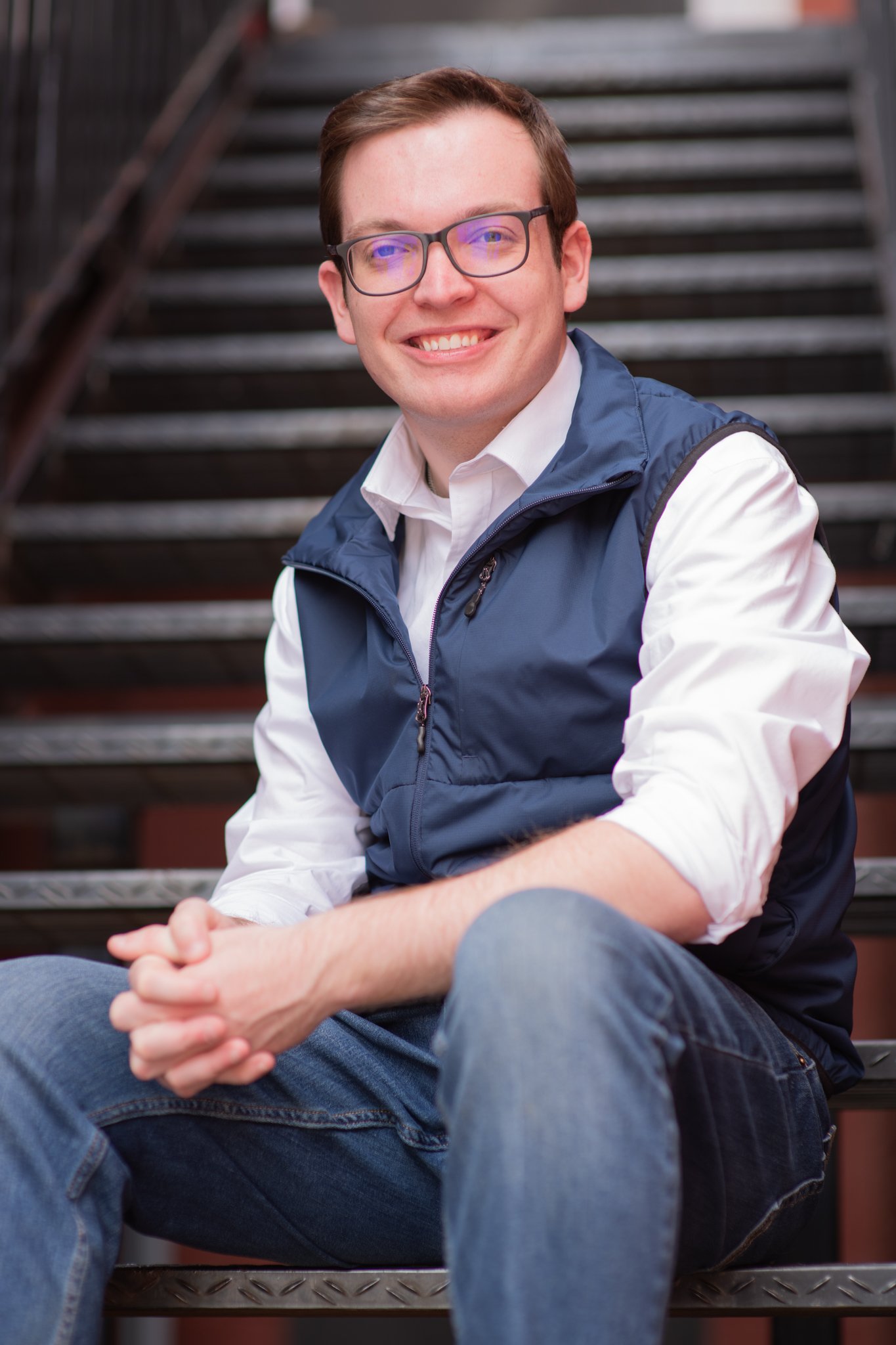 Collin McNamara, a white male, sitting on stairs, wearing a white shirt and a blue vest. He is a candidate for State Senate District 24.