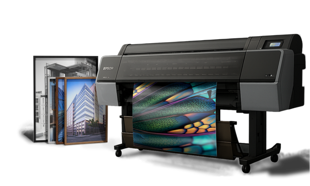 Large industrial inkjet printer printing colorful images, with framed photographs of buildings nearby.